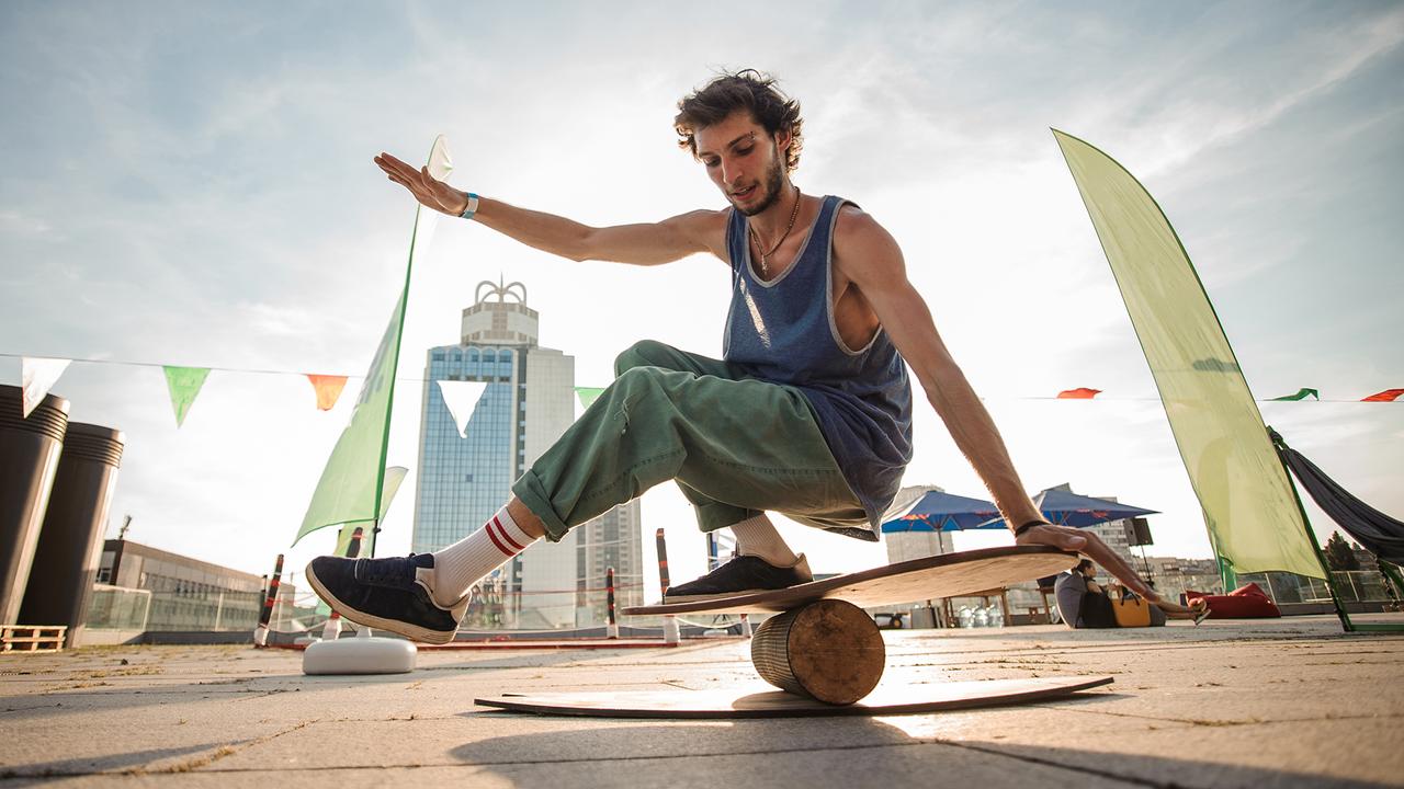 Young active man keeping balance on the wooden board against the background of city buildings on summer day - Image.