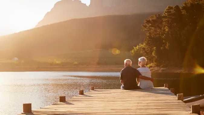 Romantic Senior Couple Sitting On Wooden Jetty By Lake.
