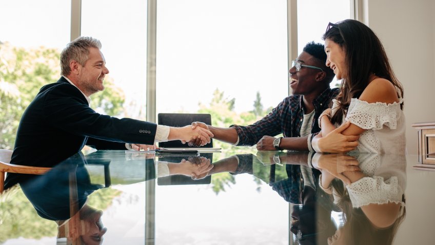 Male real estate broker shaking hands with new property owners while sitting across a table.