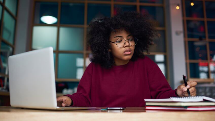Concentrated young female student in glasses learning in university campus using laptop computer and wireless connection to internet preparing to examination, attractive hipster girl writing essay.