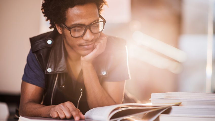 Young African American man reading a book and studying in a library.