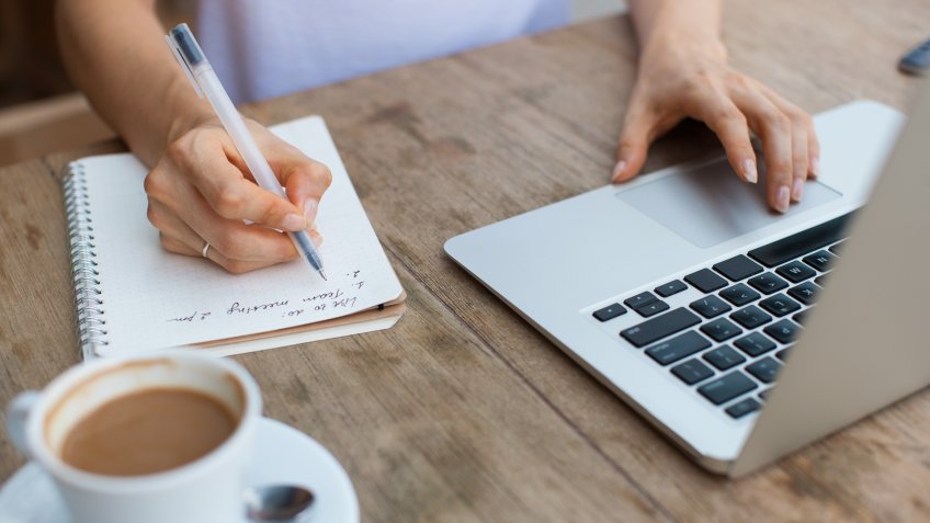 Cropped view of business woman hands working, typing on laptop computer and making notes in notebook at cafe table.