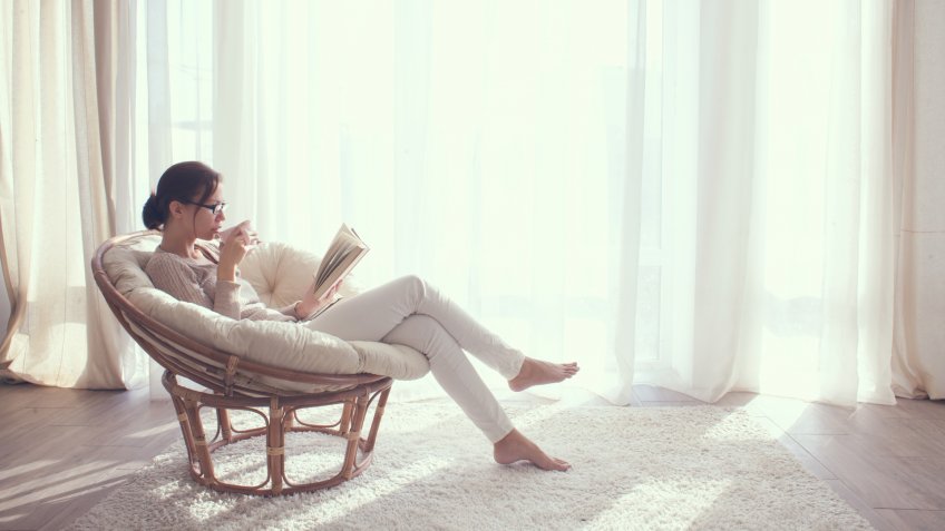 Young woman at home sitting on modern chair in front of window relaxing in her living room reading book and drinking coffee or tea - Image.