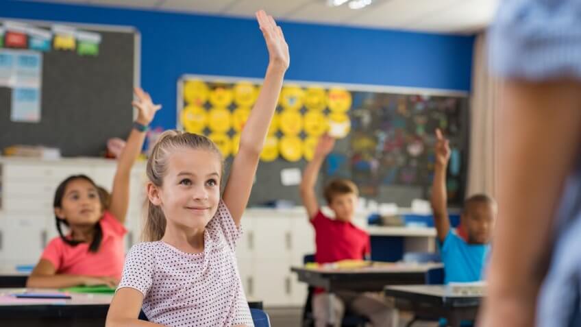 Multiethnic group of young school children raising their hands to answer a question posed by the teacher.