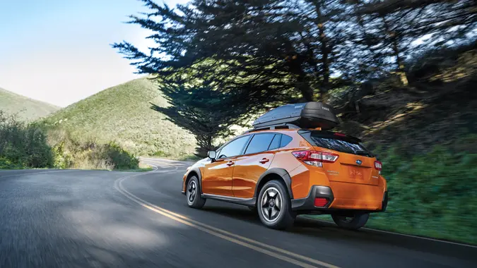 An orange Subaru Crosstrek drives on a street through green, hilly country.