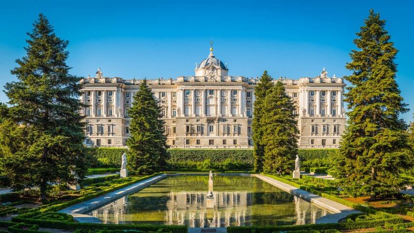The historic facade of the Palacio Real reflecting in the still waters of the ornamental pond set amongst the green trees of the Sabatini Gardens in the heart of Madrid, Spain's vibrant capital city.