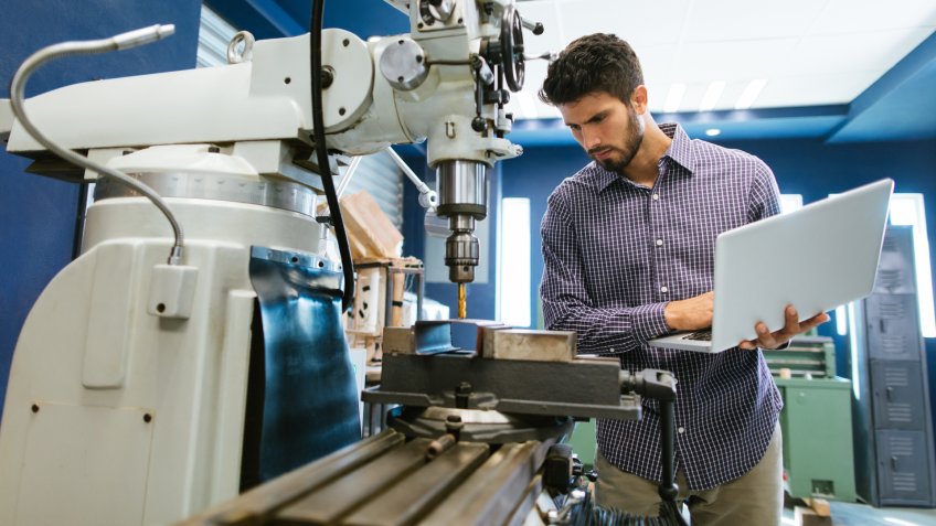 A young latin engineer calibrating a large drill with his laptop in his shop.