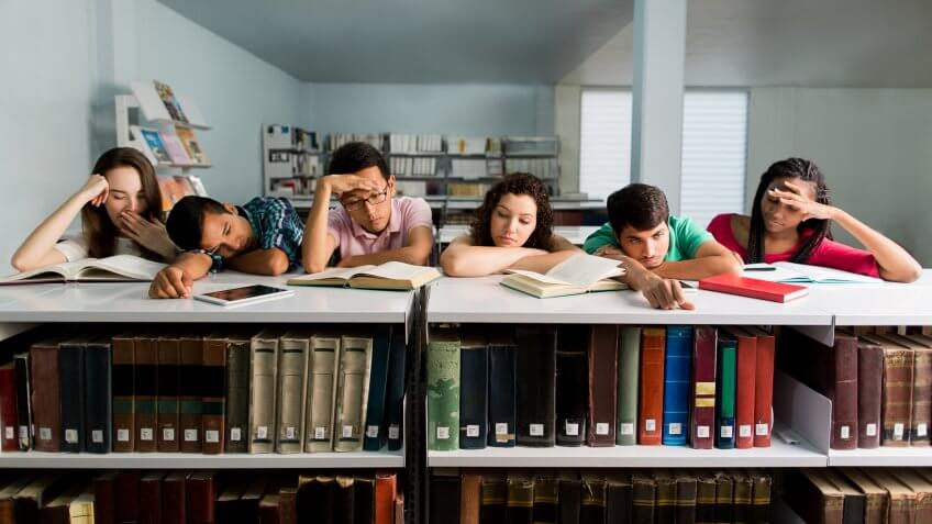 A multi-ethnic group of students leaning on a bookshelf and looking tired and sleepy in a horizontal waist up shot indoors.