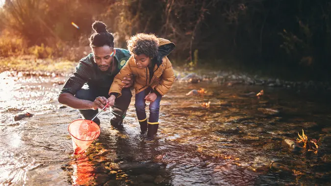 Young dad teaching son how to fish with fishing net in mountain stream at sunset.