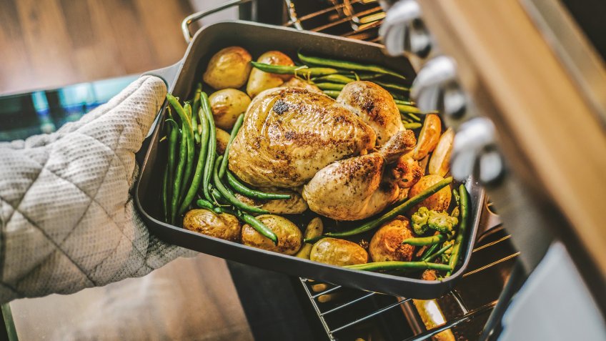 Cook taking ready fried baked chicken with vegetables from the oven.