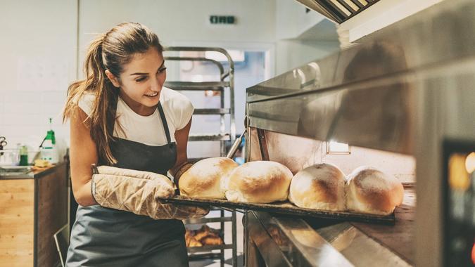 Young woman baker taking out the hot bread from the oven.