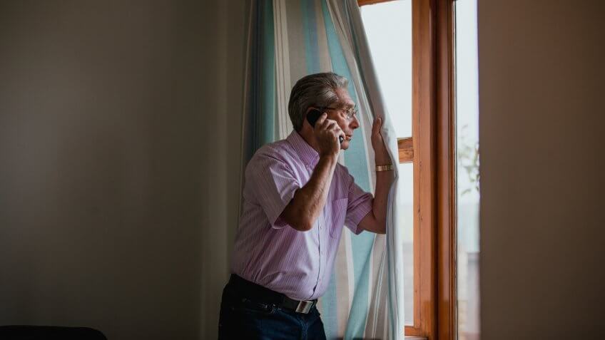A senior man uses the telephone whilst peeking out of the window.