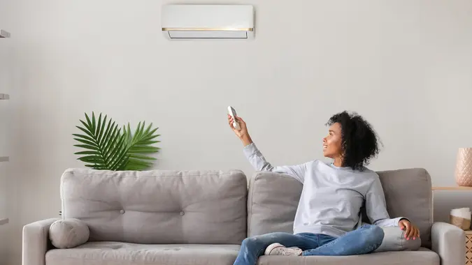 Smiling young African American woman using air conditioner, cooler system remote controller, switching, setting comfort temperature in living room, resting on cozy sofa at home, enjoy fresh air - Image.