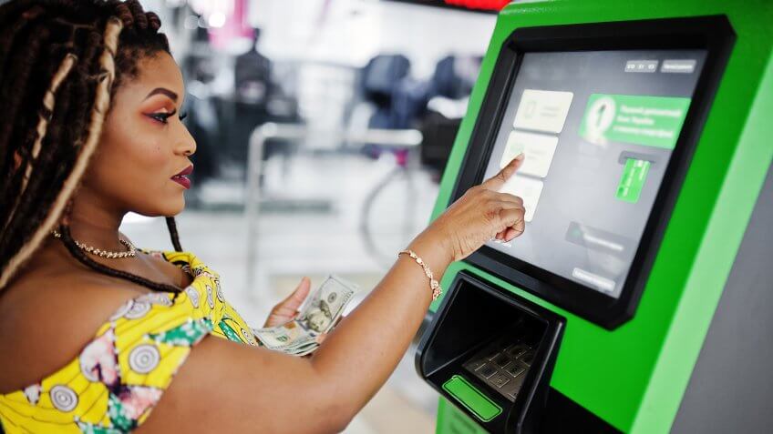 Cute small height african american girl with dreadlocks, wear at coloured yellow dress, withdrawing money dollars from credit card at ATM.