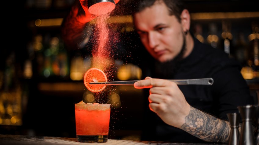 Tattooed professional bartender adding spices powder into a cocktail glass filled with a fresh strong alcoholic cocktail with slice of lemon.