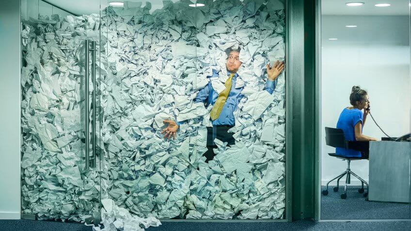A businessman is trapped in his glass office by a surplus of discarded ideas on paper .