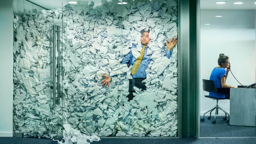 A businessman is trapped in his glass office by a surplus of discarded ideas on paper .