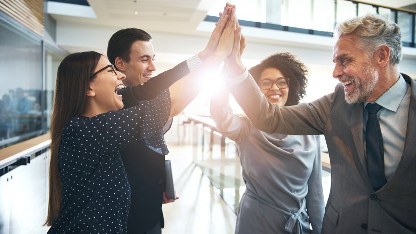 Smiling and laughing office working black and white men and women standing and giving high five to each other.