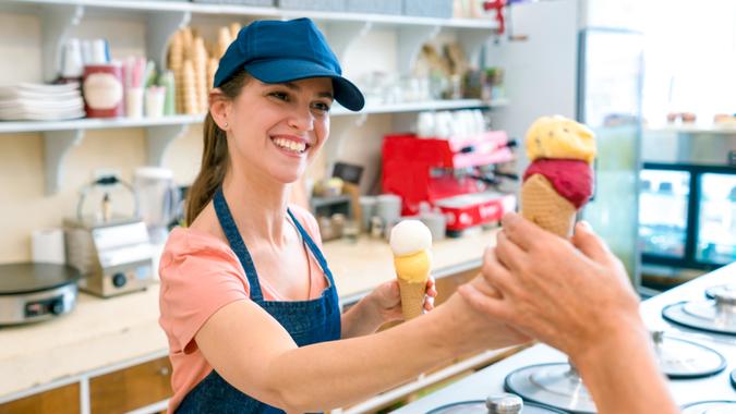 Female worker serving an ice cream to an unrecognizable customer at the ice cream parlor looking very happy and smiling.