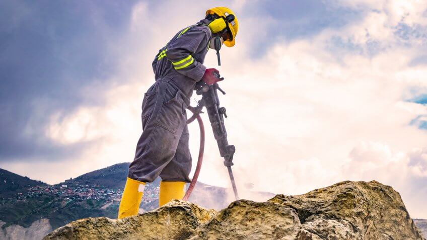 Worker with helmet and protective suit using a drilling machine on top of a large rock.