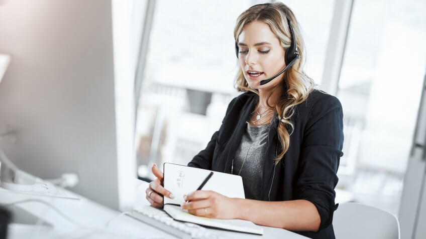 Shot of a young woman using a headset and computer at her desk in a modern office.