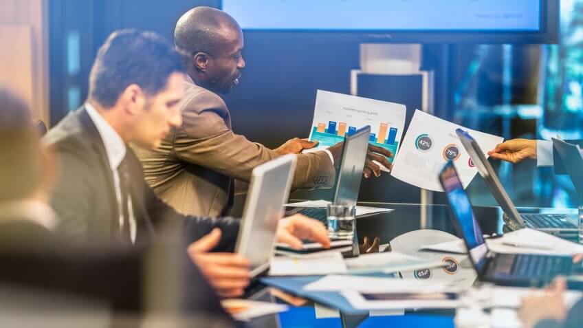 African American businessman showing infographic to his invisible colleague also holding infographic, on a board meeting at conference table.