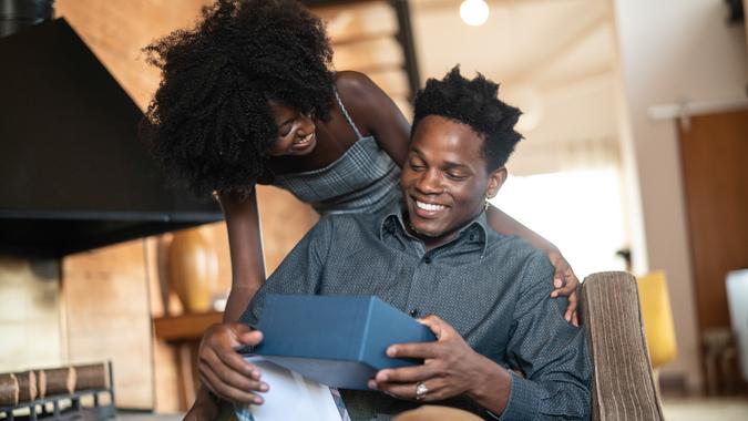 Wife making a surprise giving a present to her husband at home African descent brazilian couple exchanging gifts at home.