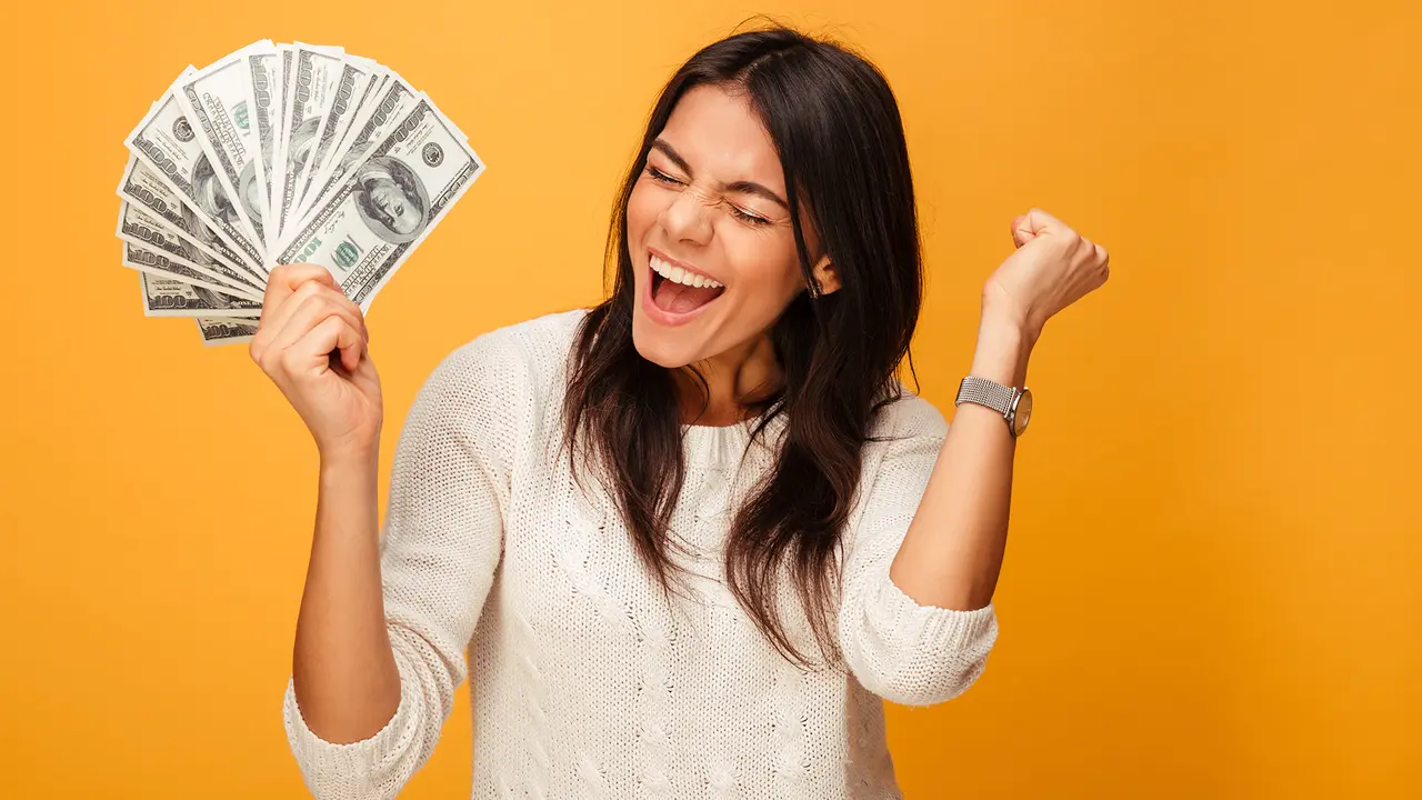 Portrait of a cheerful young woman holding money banknotes and celebrating isolated over yellow background - Image.
