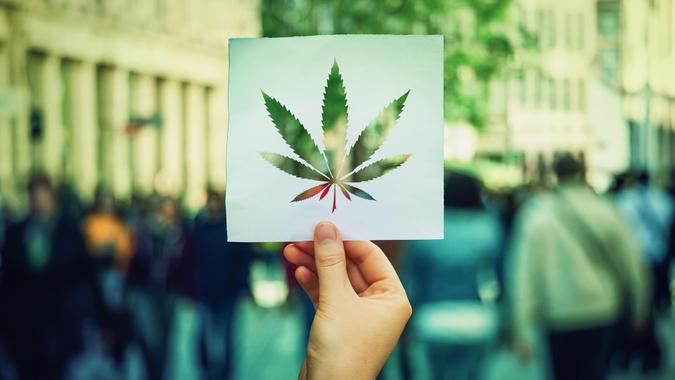 Hand holding a paper sheet with marijuana leaf symbol over a crowded street background.