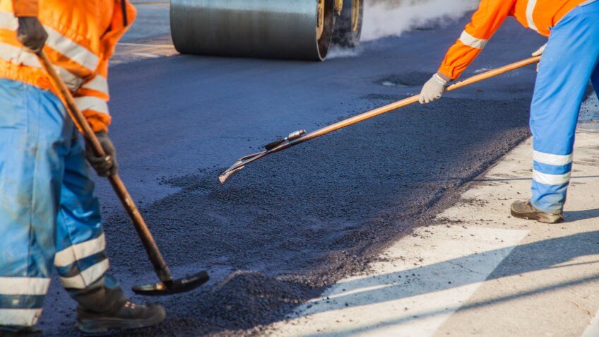 Two manual workers helping to level the asphalt surface for a steamroller to press.