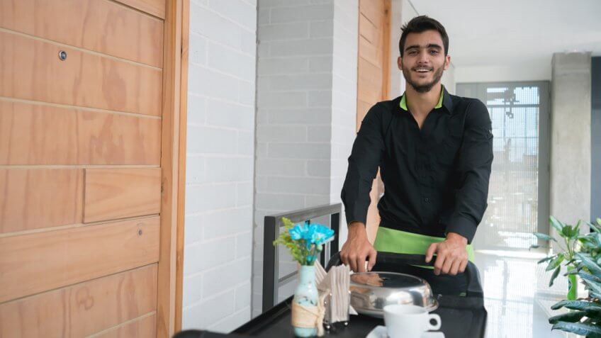 Happy man working at a hotel taking room service in a cart and looking at the camera smiling - food service concepts.