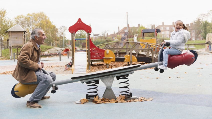Grandfather and granddaughter playing on seesaw at playground.