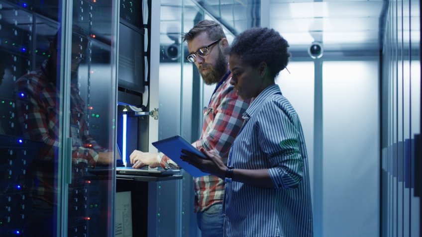Modern multiethnic man and woman with tablet using laptop in server room while checking servers.