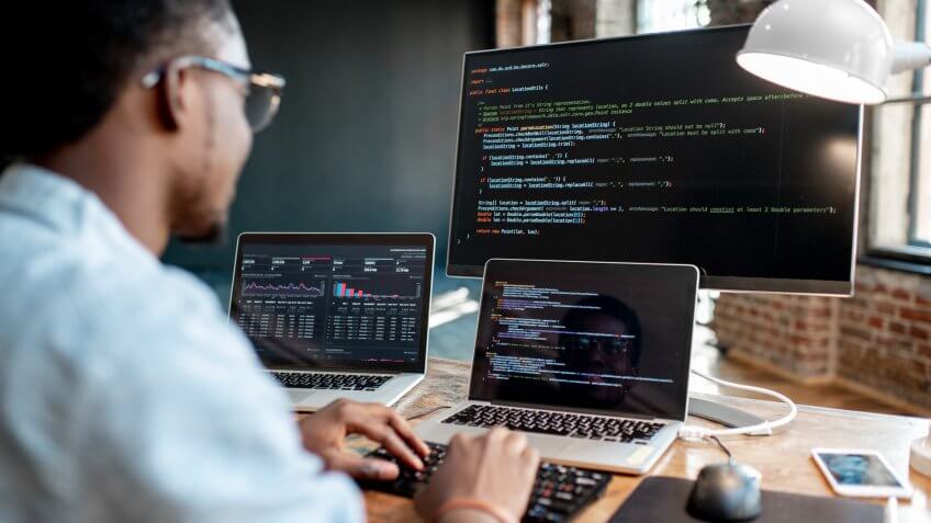 Young african male programmer writing program code sitting at the workplace with three monitors in the office.