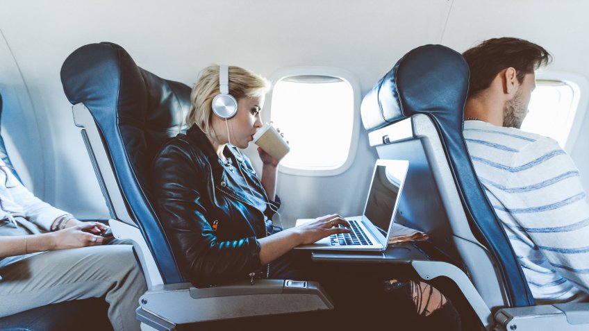 Young blonde woman sitting inside an airplane, using laptop and drinking coffee.