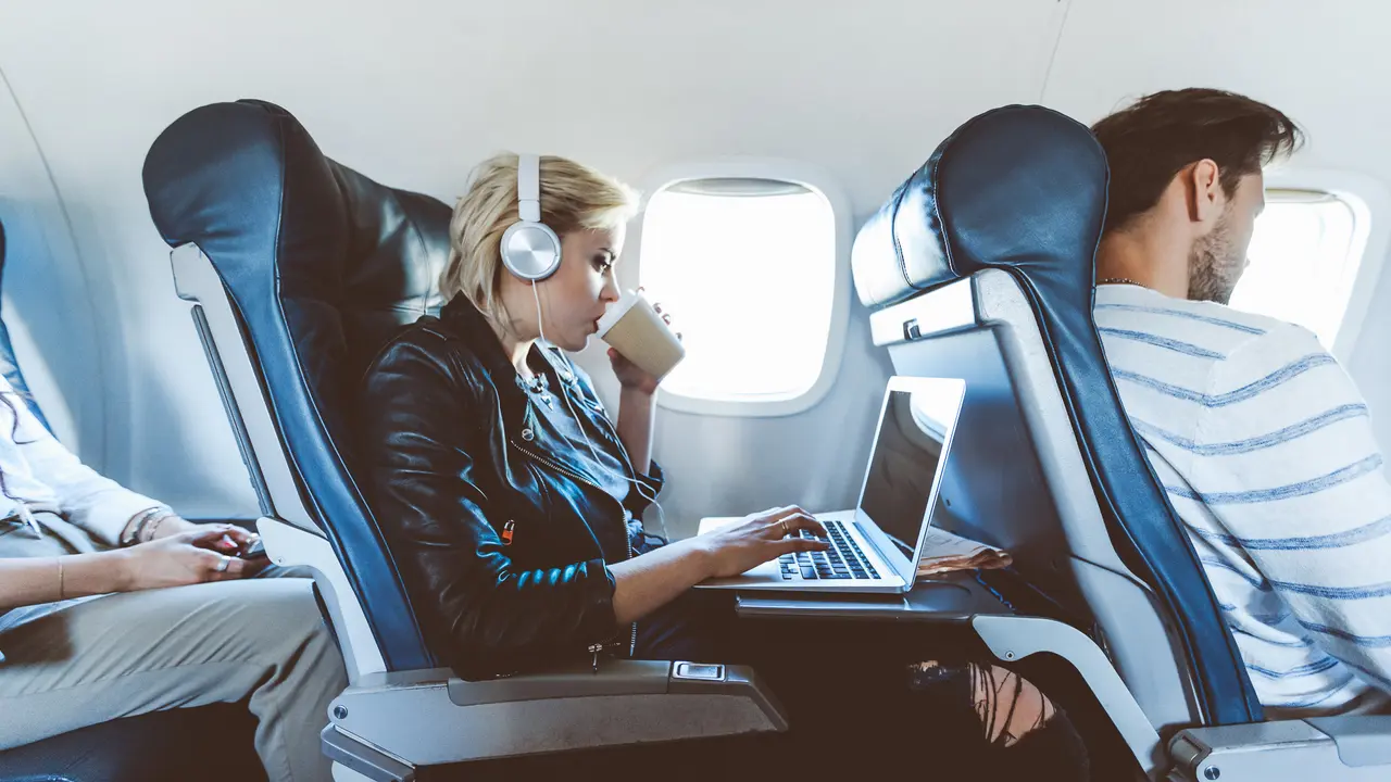 Young blonde woman sitting inside an airplane, using laptop and drinking coffee.