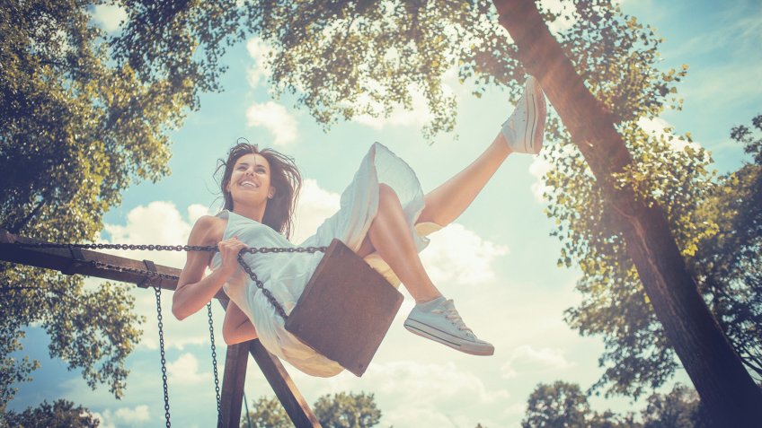 A happy young woman laughing on a bright sunny day while having fun on a swing in the park.