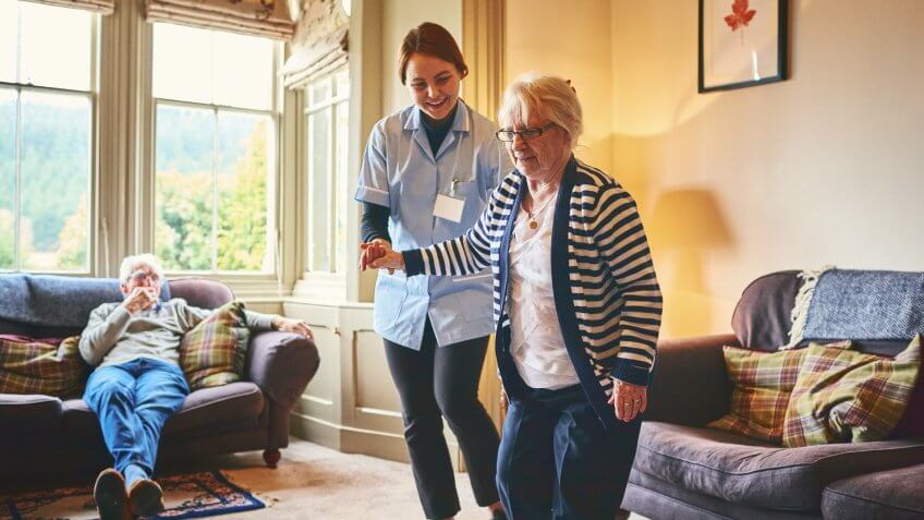 Young female nurse supporting old woman to walk indoors with senior man relaxing on sofa at back*INSPECTORS PLEASE READ*THE LEAF IMAGES ON THE WALLS ARE MADE BY THE OWNER OF THE PROPERTY SO ARE COVERED BY THE PR ATTACHED.