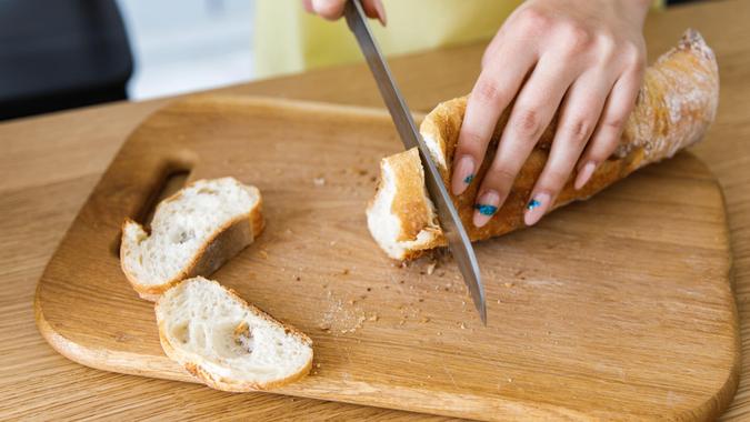 Woman slicing bread with on chopping board on wooden table close up.