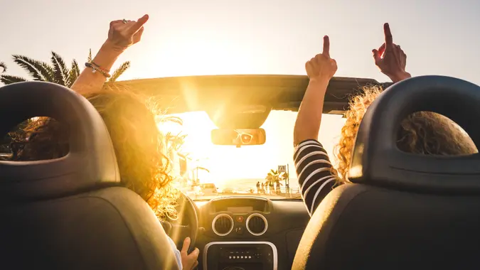 Couple of woman friends traveling and driving having a lot of fun dancing in the car with opened roof and summer vacation sunset ocean in front - concept of friendship together and nice lifestyle for independent girls.