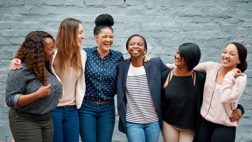 Portrait of a diverse group of young women standing together against a gray wall outside.
