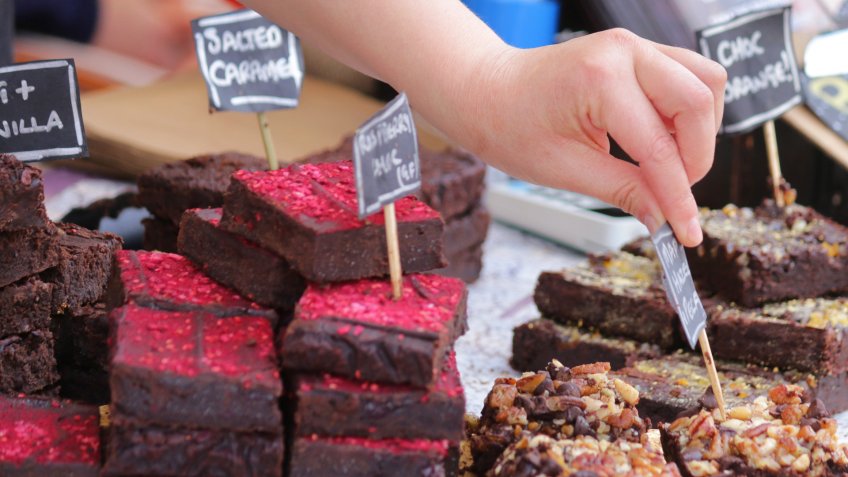 Stock photo of homemade chocolate brownies recipe tray bake, home baking cakes cut into squares, baked and topped with toffee and peanuts, chopped nuts, raspberries, raspberry powder, salted caramel and vanilla, stack pile at bakery stall in street food market.