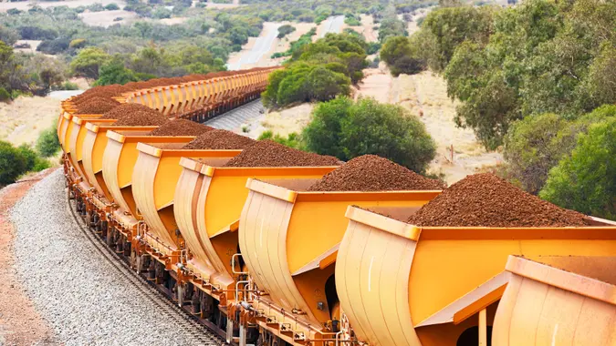 Heavy train loaded with brown hematite iron ore in orange freight wagons (cars) passes through rolling hills.