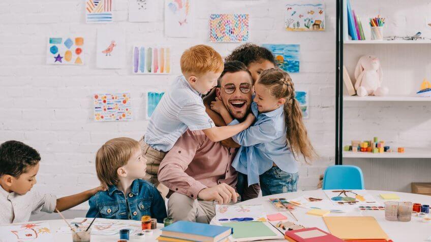 interracial kids hugging happy teacher at table in classroom.