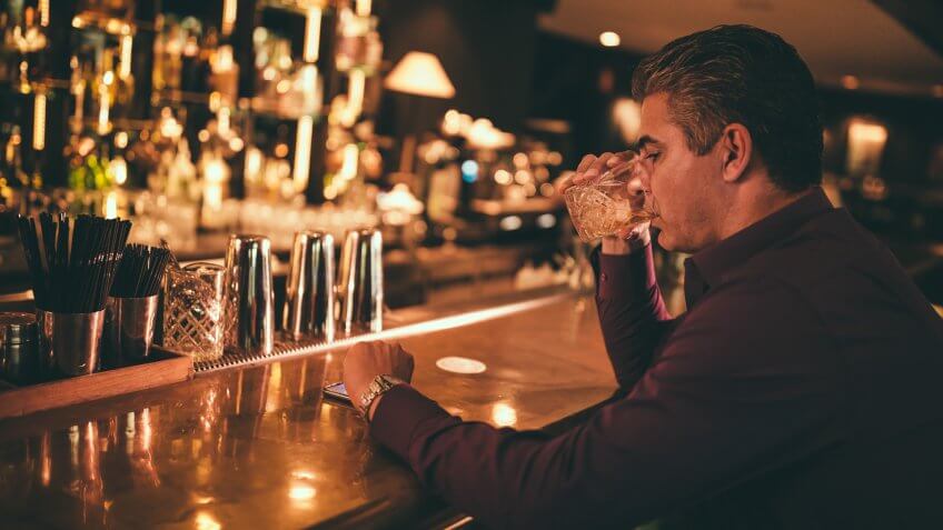 Elegant senior man sitting on bar counter drinking whiskey and looking at watch.