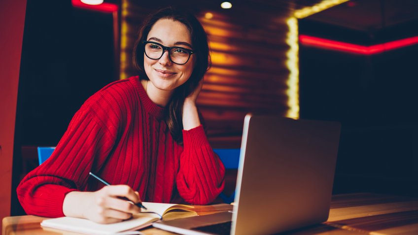 Smiling young woman enjoying freelance work in cafe interior making notes of ideas in notepad, positive female student doing homework in good mood satisfied with wifi for learning on laptop computer - Image.