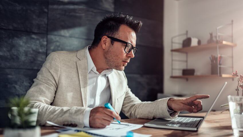 Businessman wearing linen suit and eyeglasses sitting at his desk using laptop and highlighting text with blue marker in his office.