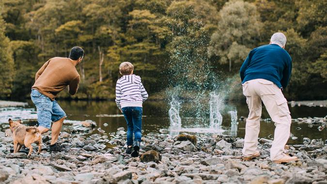 Little boy is skimming pebbles on a lake with his father and grandfather.