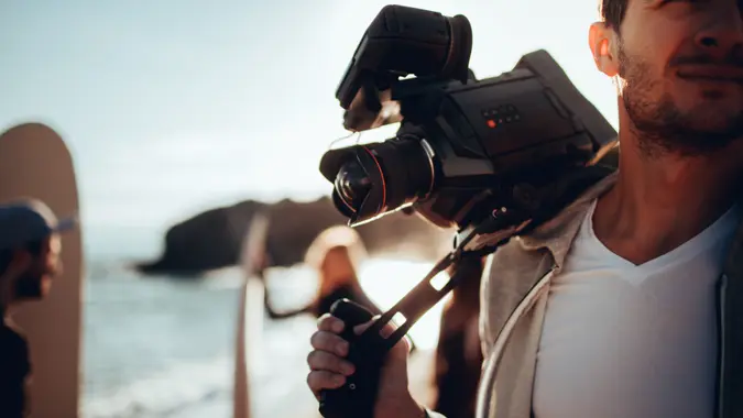 Young man holding video camera, having a photo shoot with his friends at the beach, while they are preparing to surf.