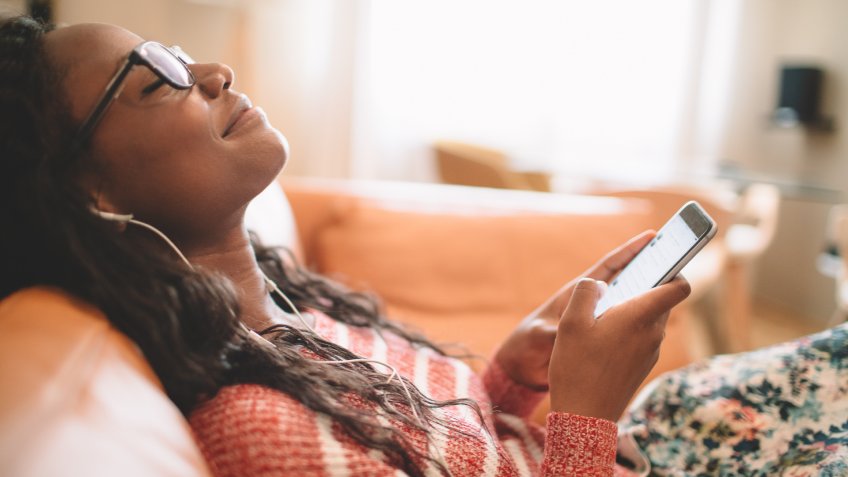 Photo of a smiling young woman using her mobile phone while resting on the couch in her living room.